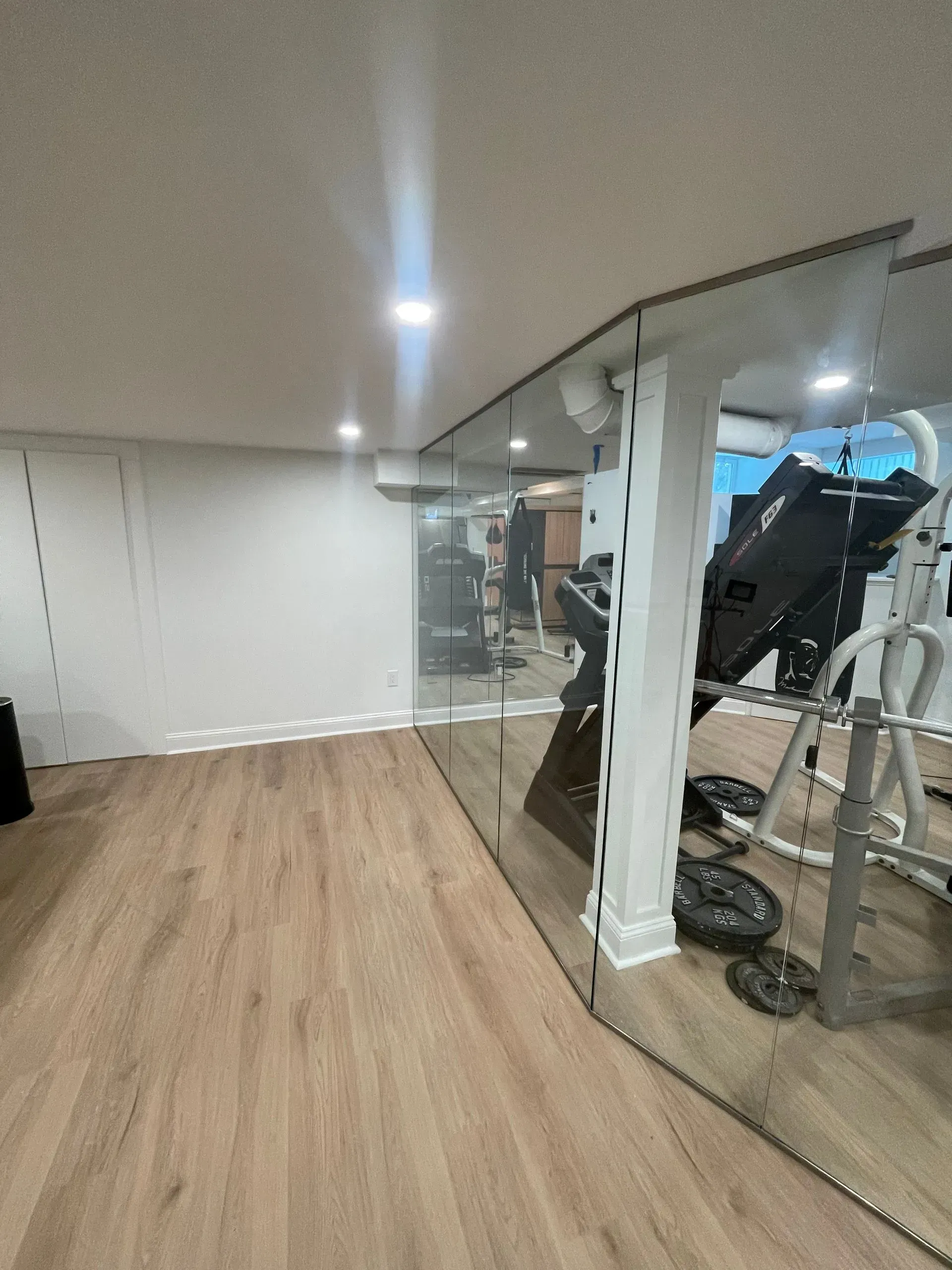 A home gym featuring light wood-look flooring, white walls, and a large wall-to-wall mirror reflecting fitness equipment.