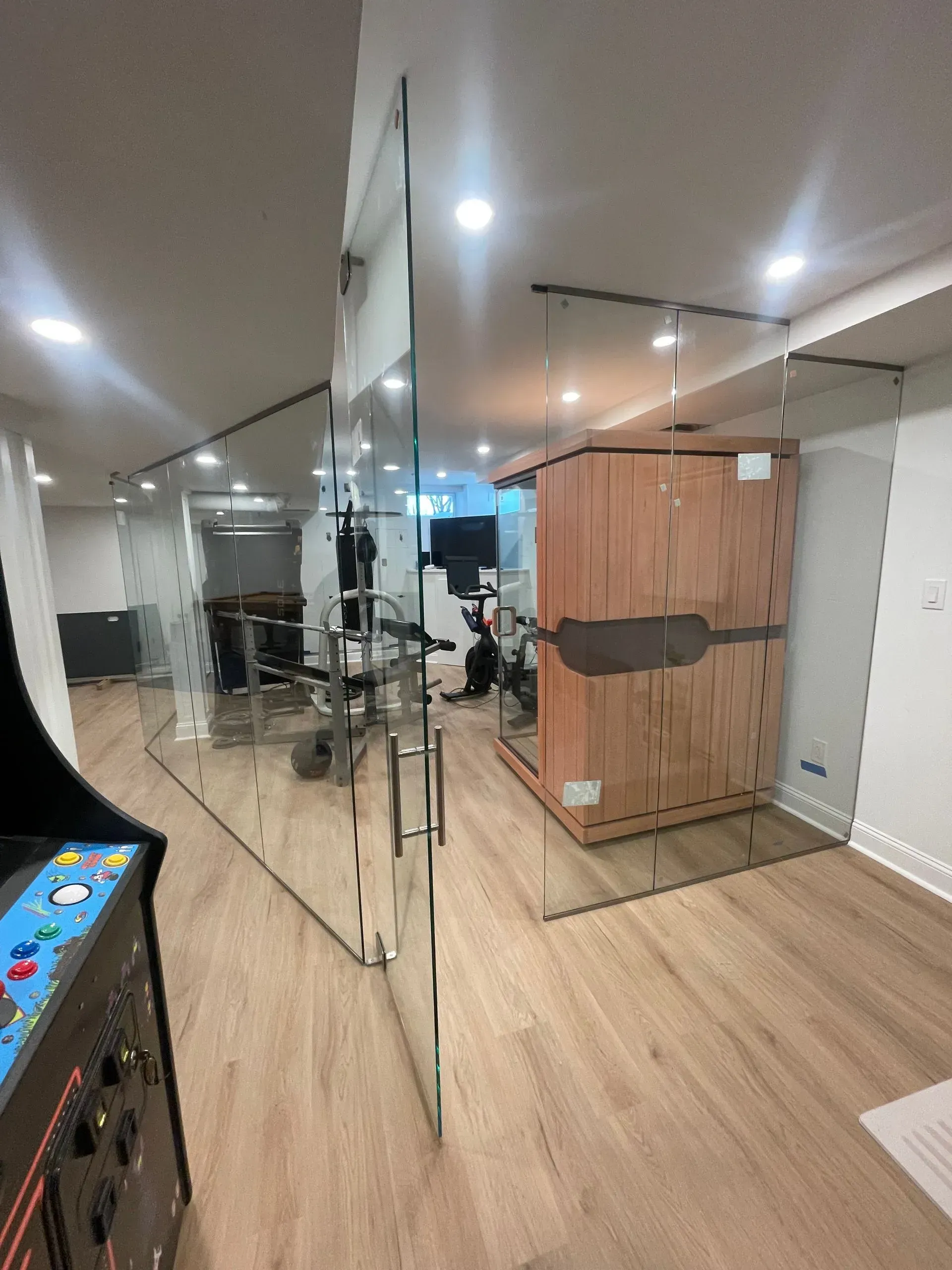 A wooden sauna stands in a basement home gym separated by glass walls, with an arcade machine in the foreground.