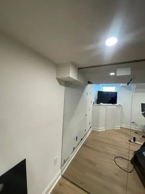 An unfinished white-walled basement room with light wood flooring, a ceiling soffit, and a television in the background.