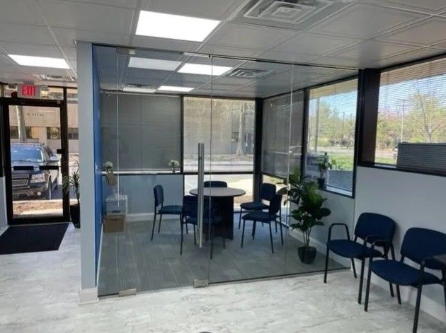 An office waiting area featuring a glass-enclosed meeting room with a table, chairs, and a plant, plus rows of chairs.