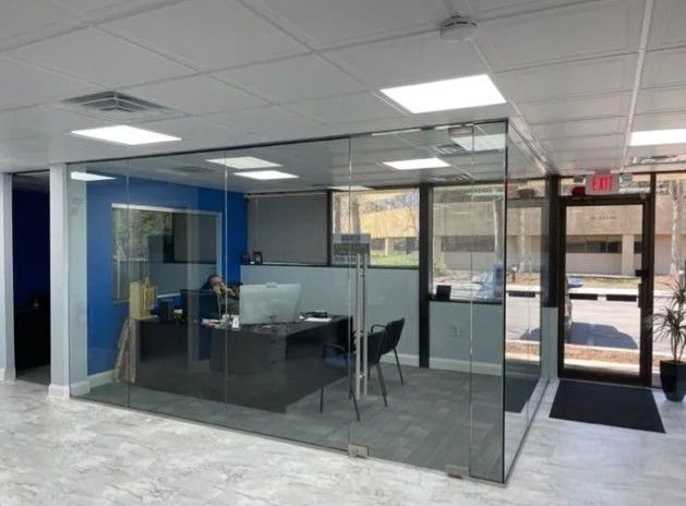 Modern glass-walled office space featuring a desk, computer, and chair, positioned beside a doorway and blue wall.