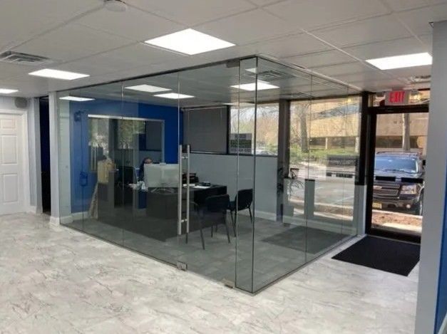 An indoor, glass-walled office pod with a desk and chairs, situated in a lobby with tile floors and a nearby exit door.