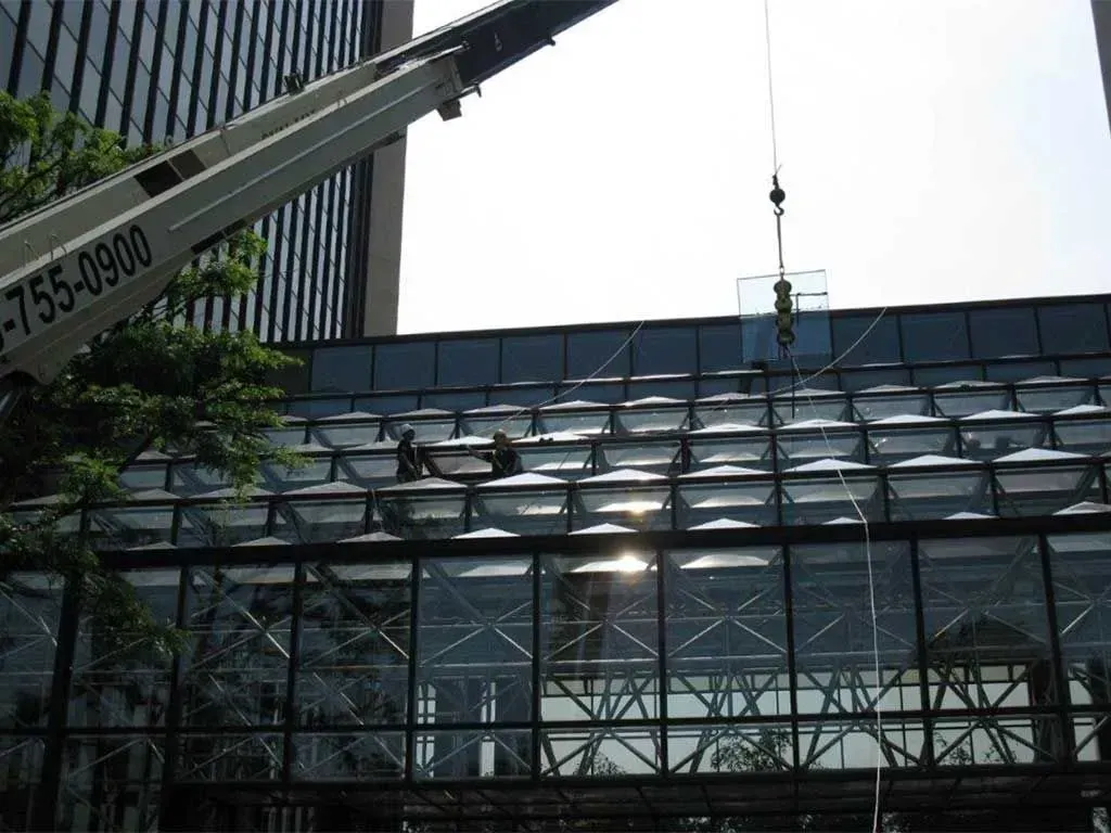 A crane lowers a large glass panel onto the sloped roof of a building as workers guide the installation from above.