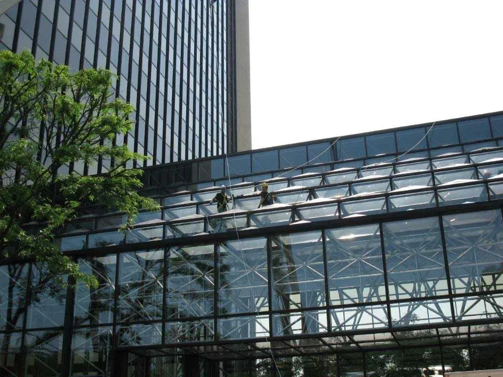 Two window cleaners working on the glass roof of a modern, multi-story office building.