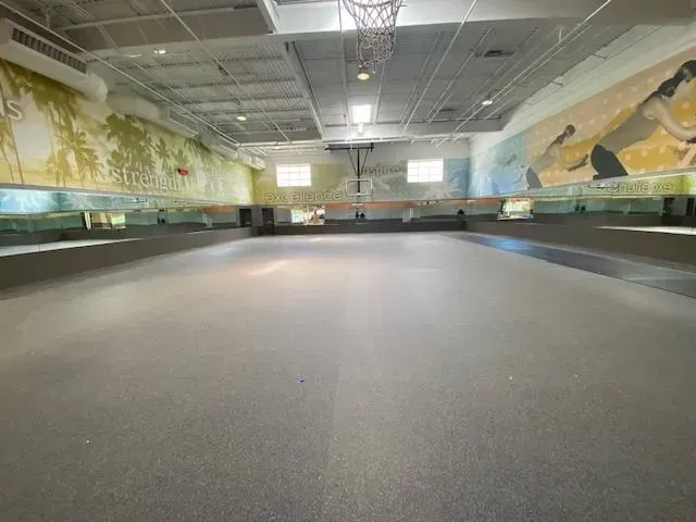 An empty indoor gym with gray rubber flooring, mirrors along the walls, a basketball hoop, and painted wall murals.