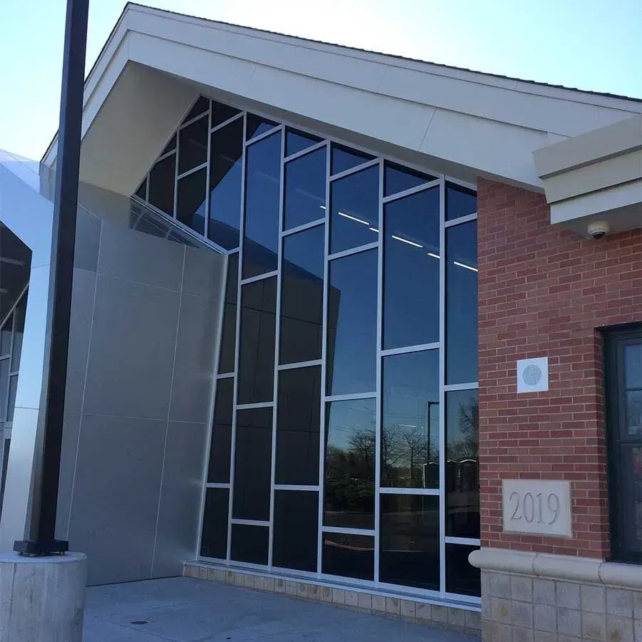 Modern building entrance featuring a large, irregularly partitioned glass window wall, brick facade, and a 2019 plaque.
