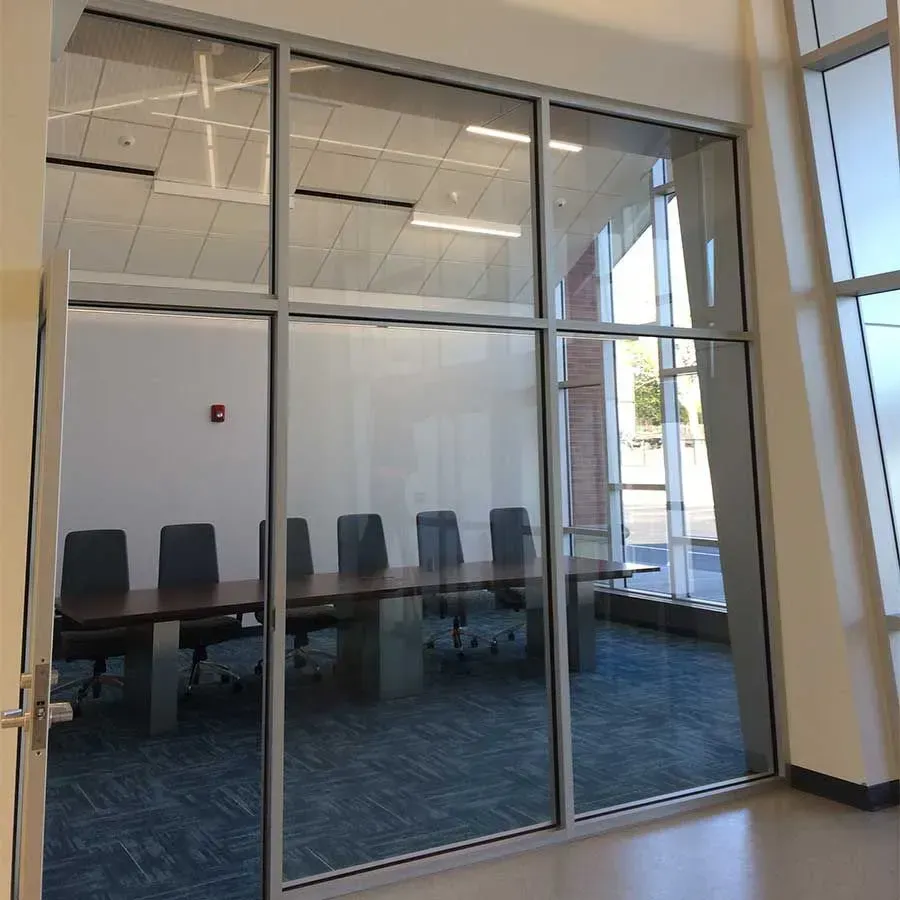 A glass-walled conference room with a long table and chairs set against a white wall with a red fire alarm.