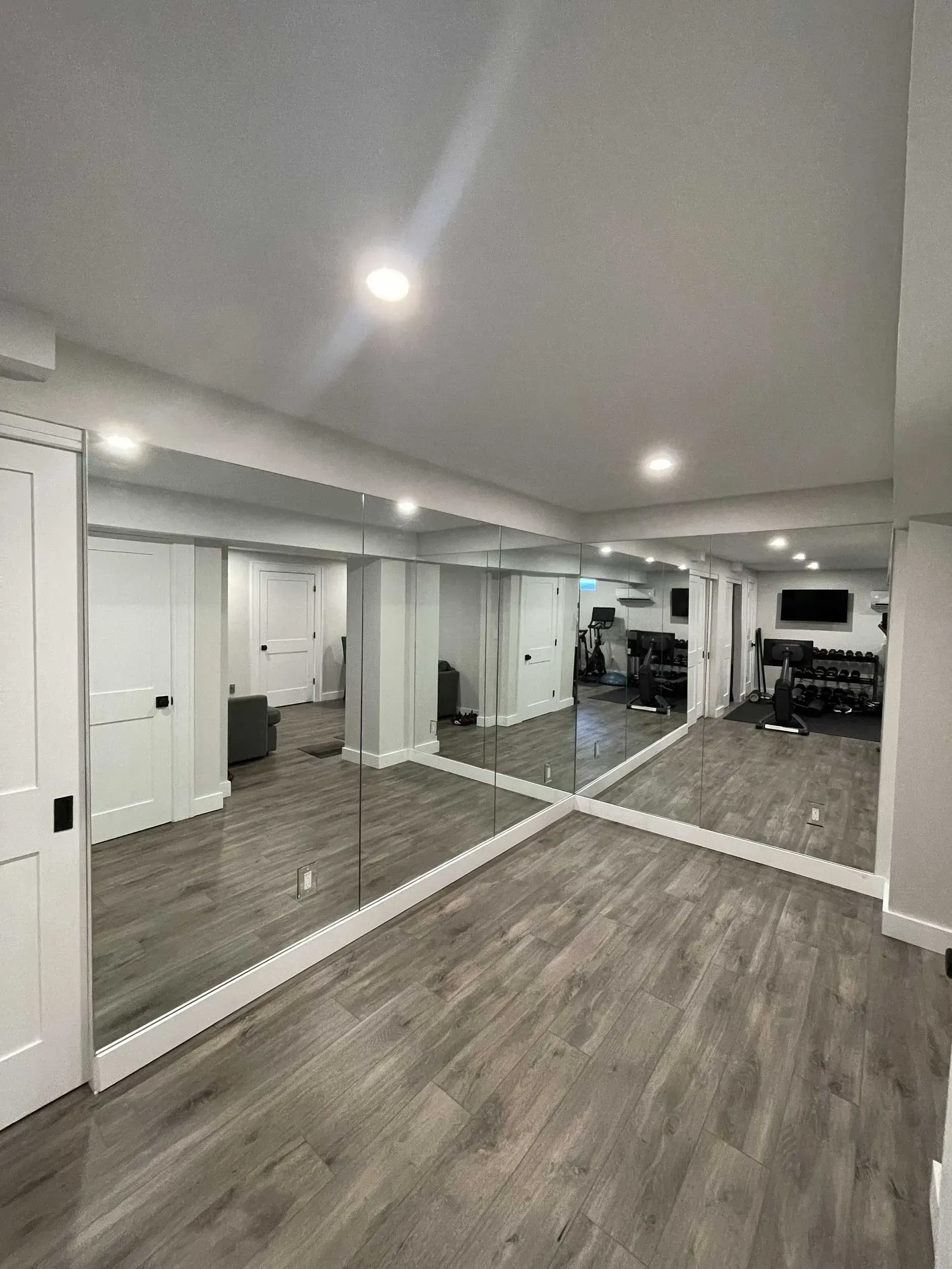 A basement room with grey wood-look flooring and a wall of large mirrors reflecting the space.