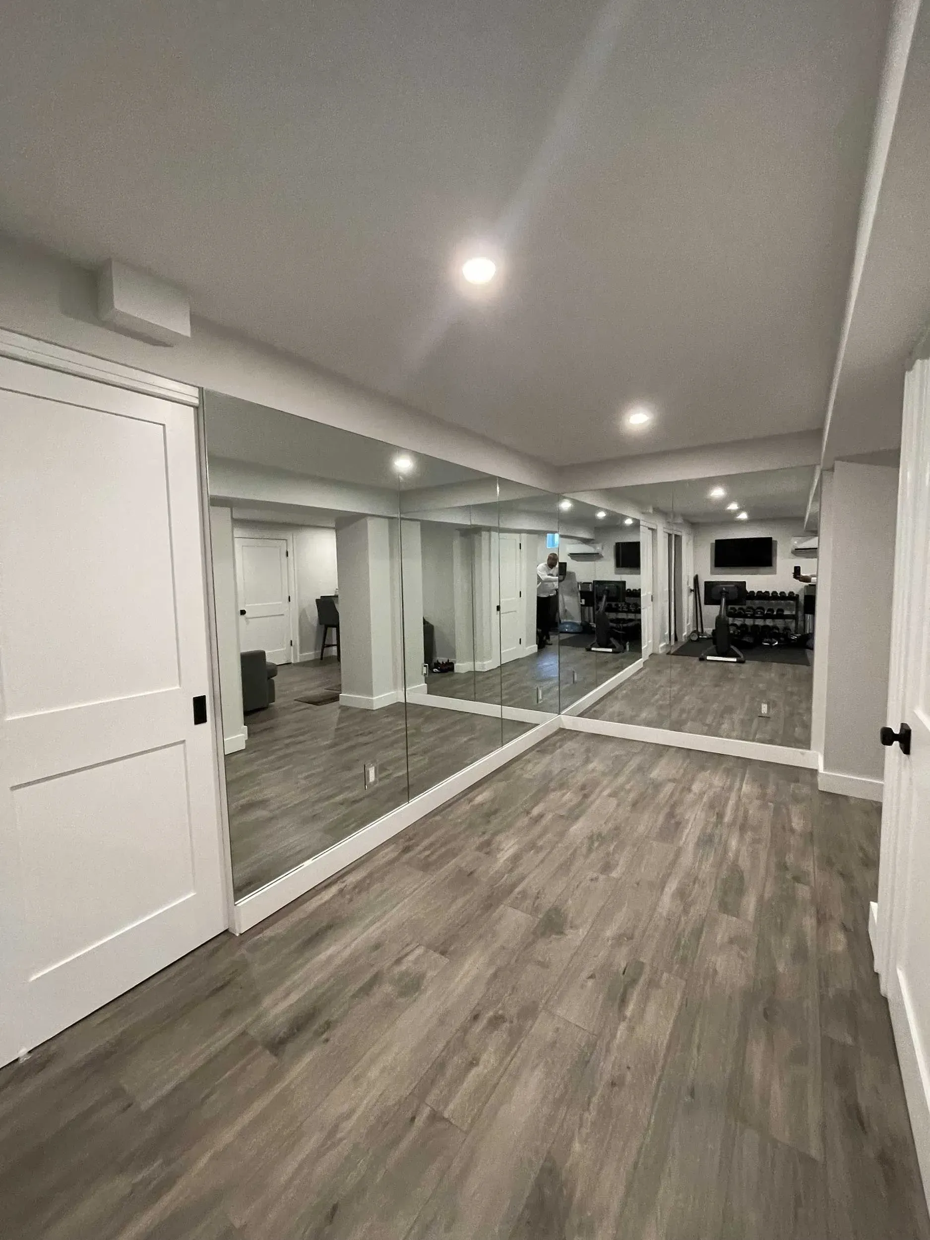 A finished basement with light gray wood flooring, white doors, and a large mirror covering the back wall.