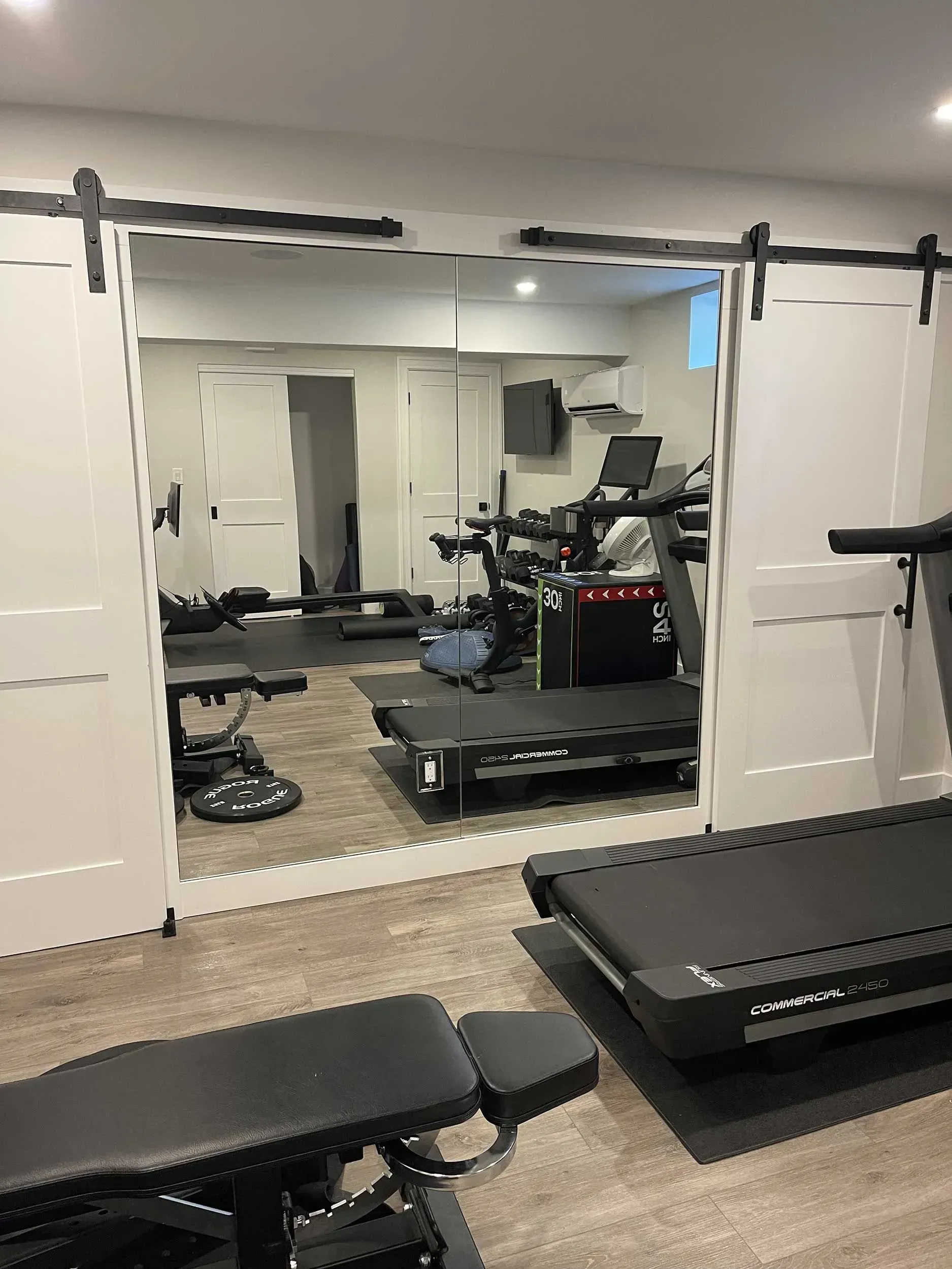 A home gym with a treadmill, workout bench, and wall-to-wall mirrors framed by sliding white barn doors.