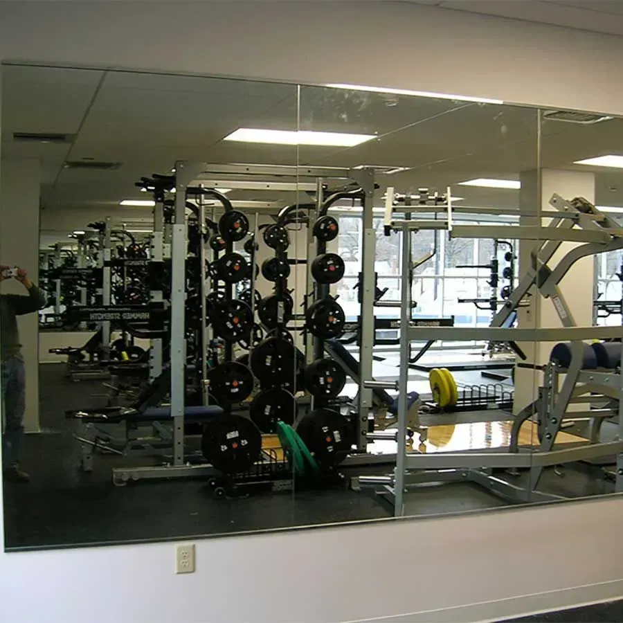 A person stands in front of a large mirror in a gym, reflecting various weight racks, equipment, and weight plates.