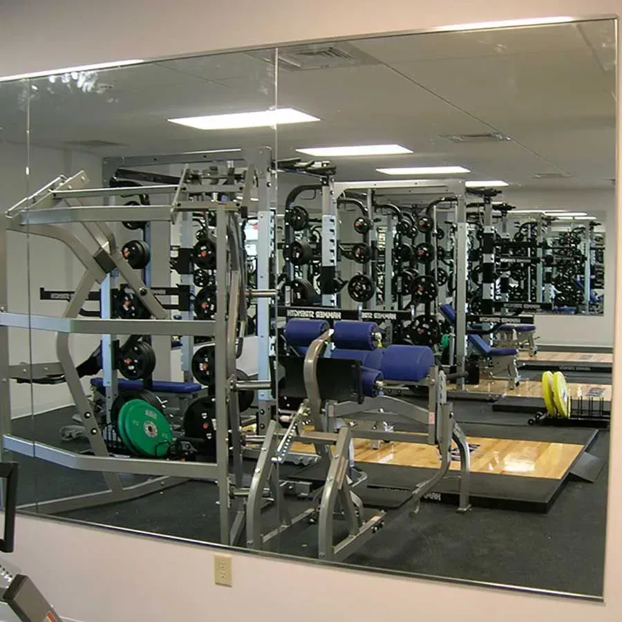 A large gym mirror reflects multiple weightlifting racks, barbell plates, and exercise benches in a training room.