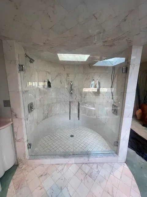 A curved glass shower enclosure with marble tile walls and floors, featuring dual glass doors and a central showerhead.