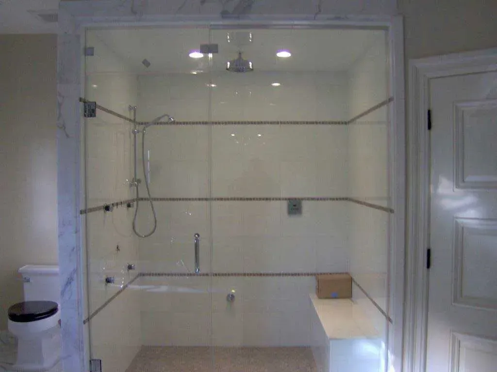 A glass-enclosed shower featuring white tiled walls, marble trim, a rainfall showerhead, and a built-in bench.