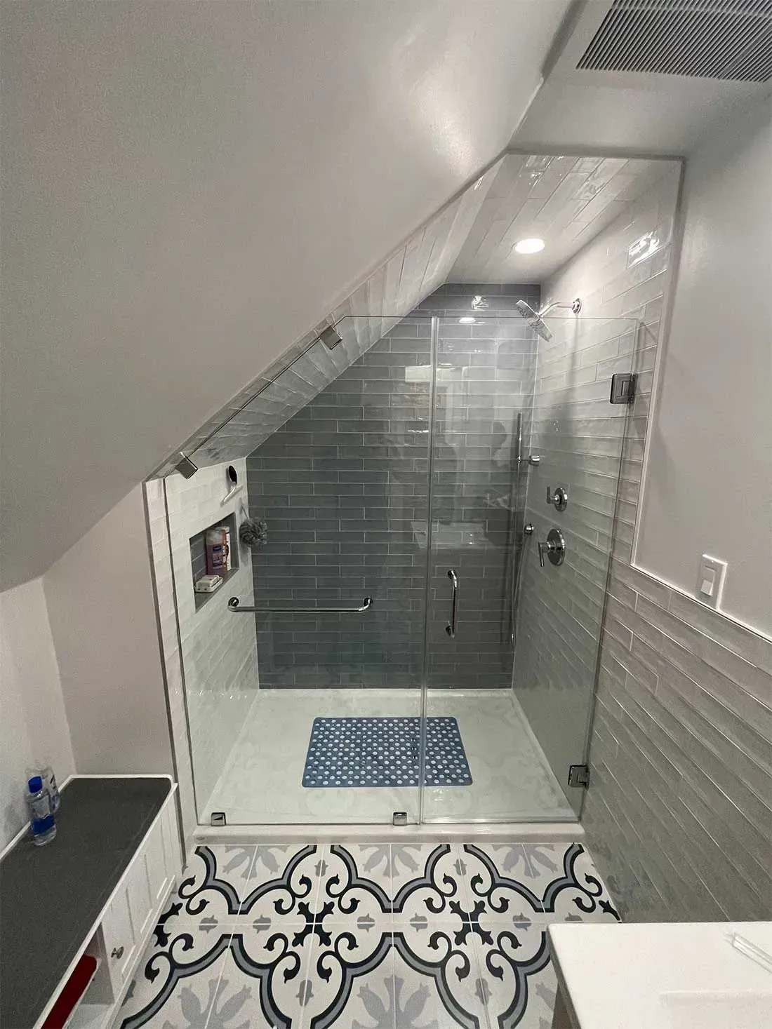 A glass-enclosed shower featuring dark gray tiled walls, a built-in niche, and patterned flooring in a slanted attic space.