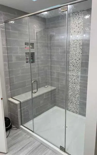 A walk-in shower with a glass door, light grey brick-style tiles, a built-in bench, and a vertical mosaic accent strip.