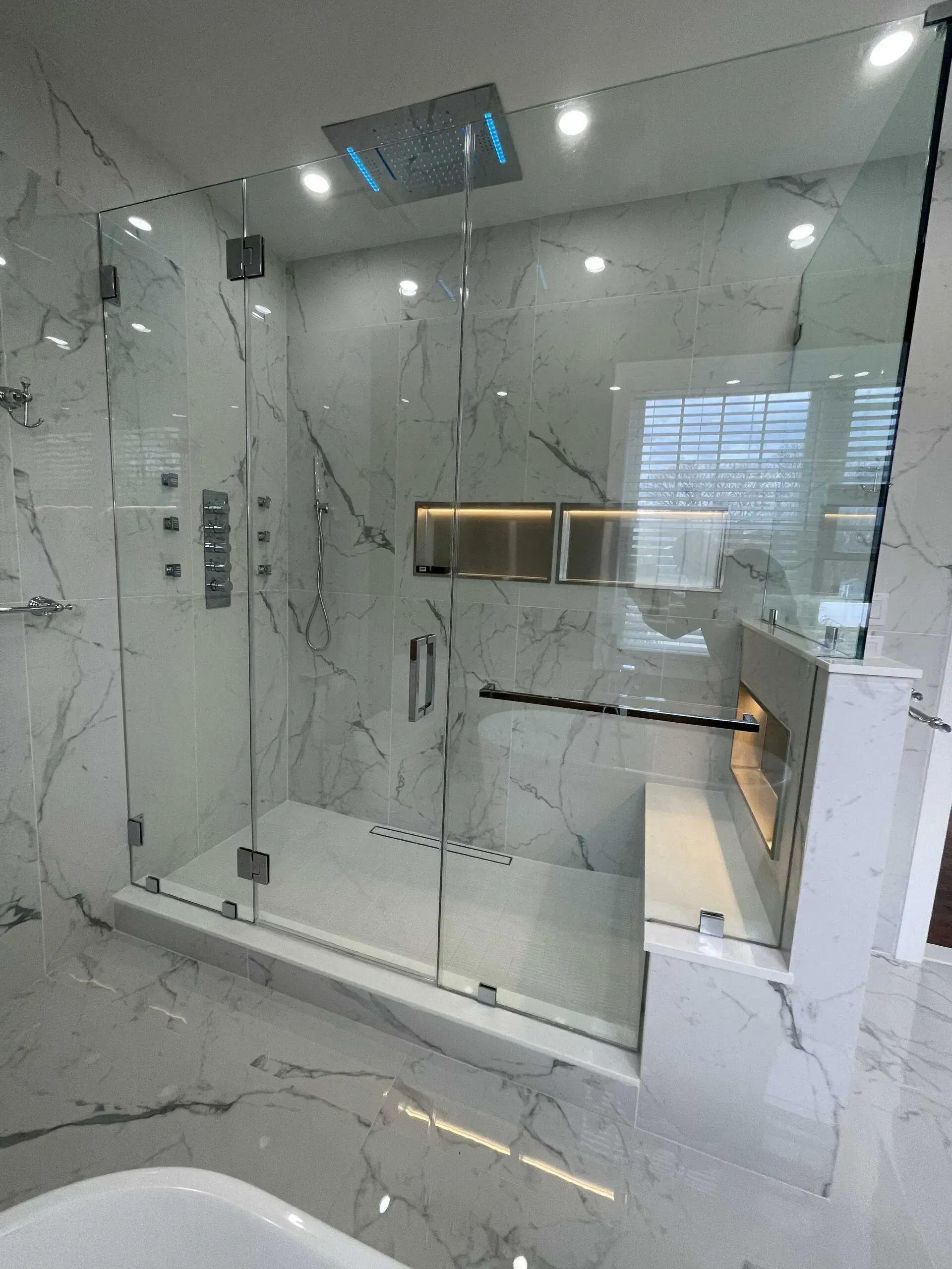 A modern glass-enclosed shower with white marble walls, a rainfall showerhead, and a built-in bench with a grab bar.