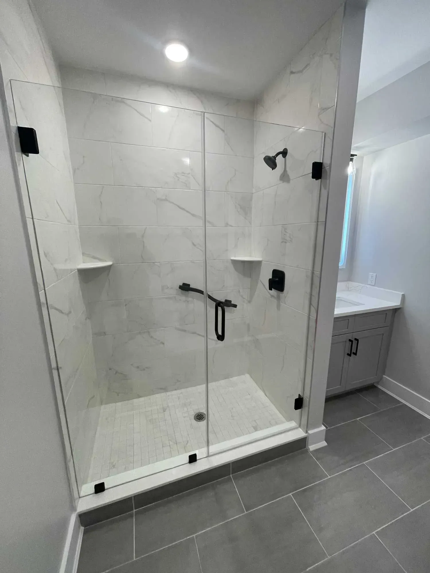 A walk-in shower with white marble-patterned tiles, a glass door, black hardware, and two built-in corner shelves.