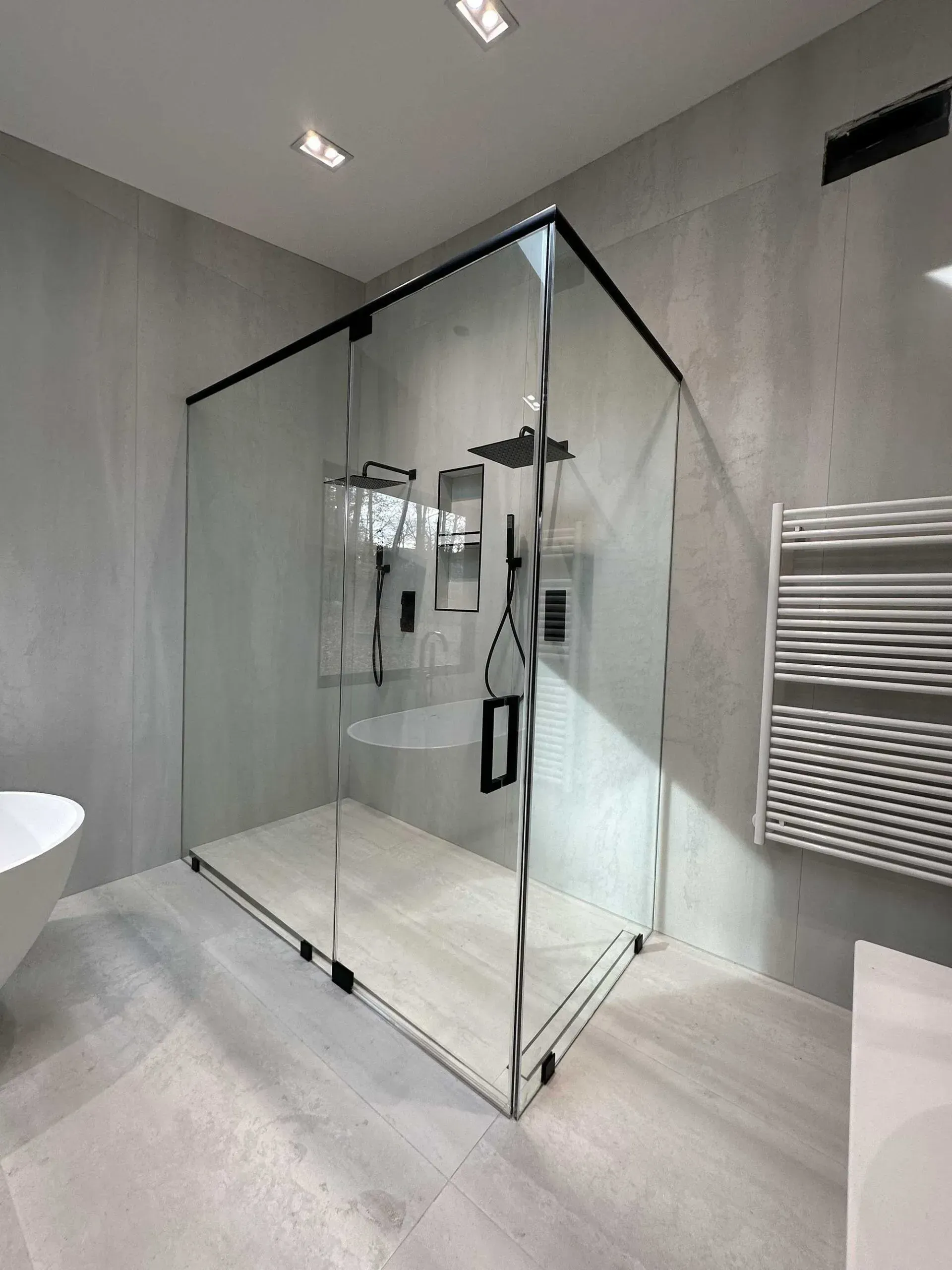 A modern, minimalist glass walk-in shower with black hardware in a light-gray tiled bathroom.