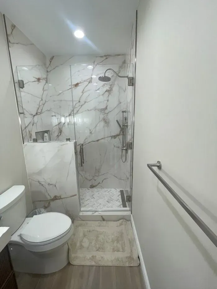 A white toilet sits next to a glass-enclosed shower with marble-patterned walls and a stone floor mat.