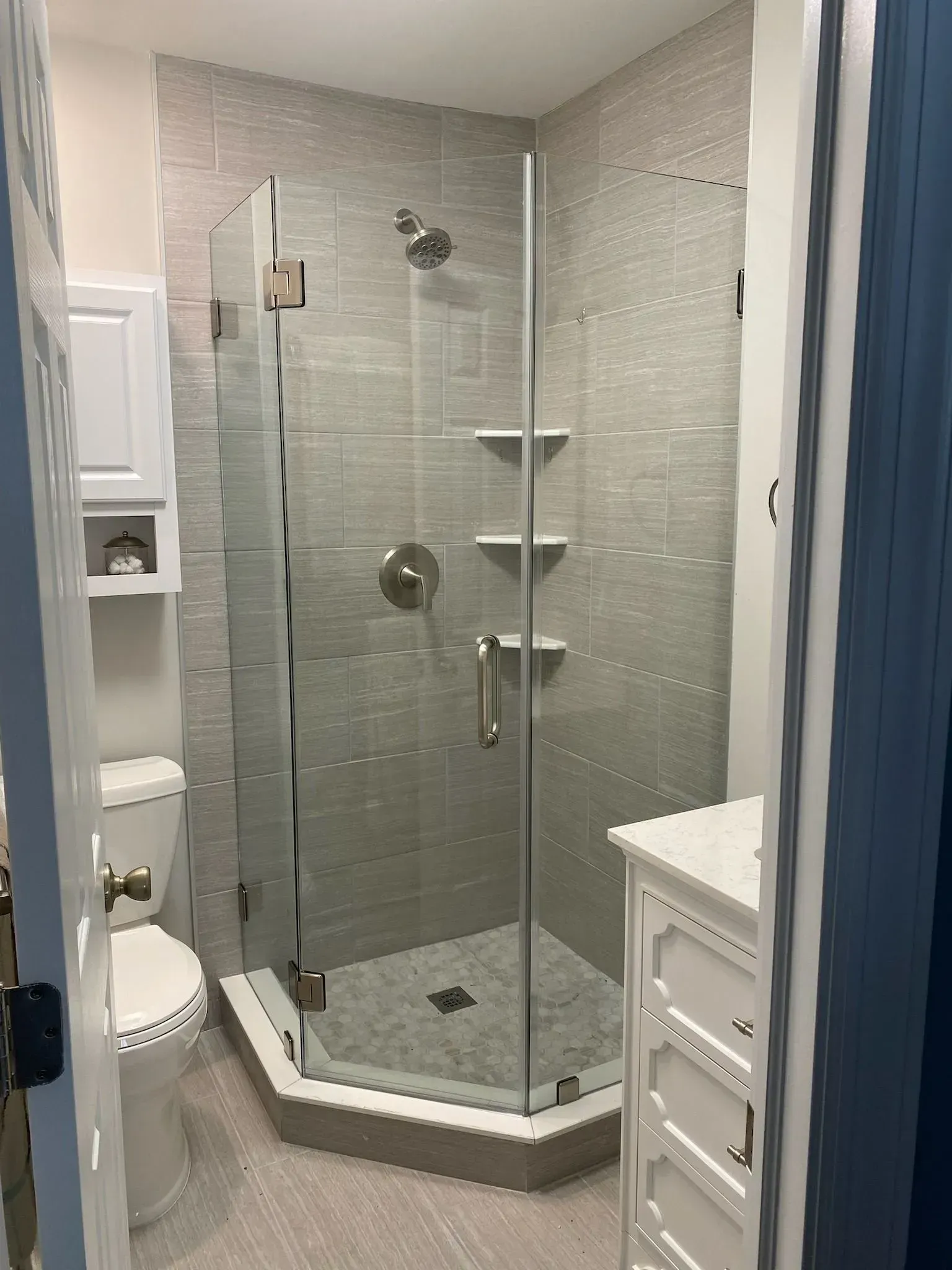 A bathroom featuring a neo-angle glass shower with grey tile, white cabinets, and a white toilet.