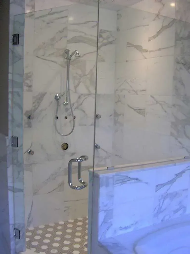 A glass-enclosed shower with white marble-patterned tile walls and light-colored hexagonal floor tiles.