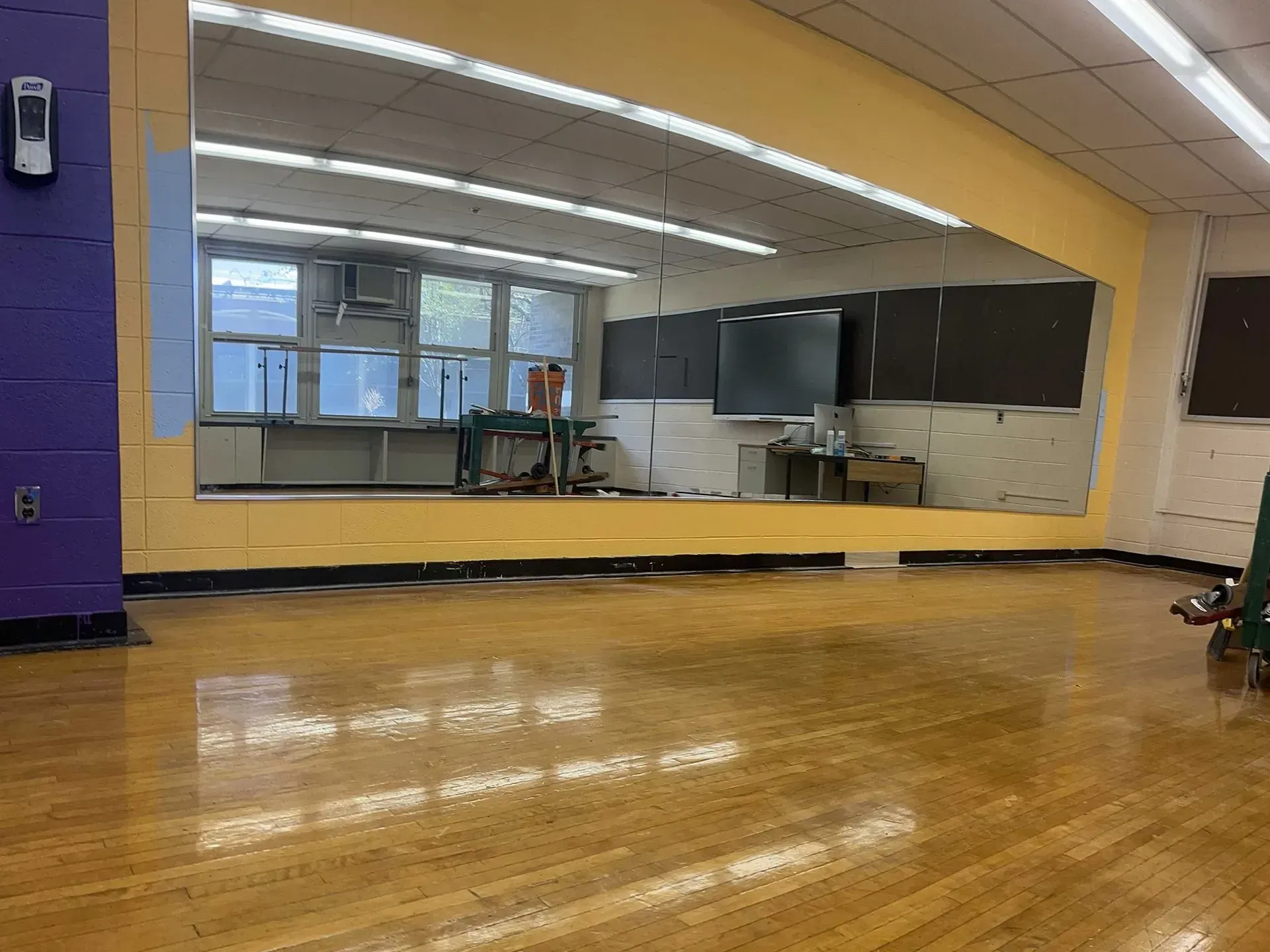 A large empty dance studio with polished wooden floors, a wall-to-wall mirror, and a purple pillar on the left.