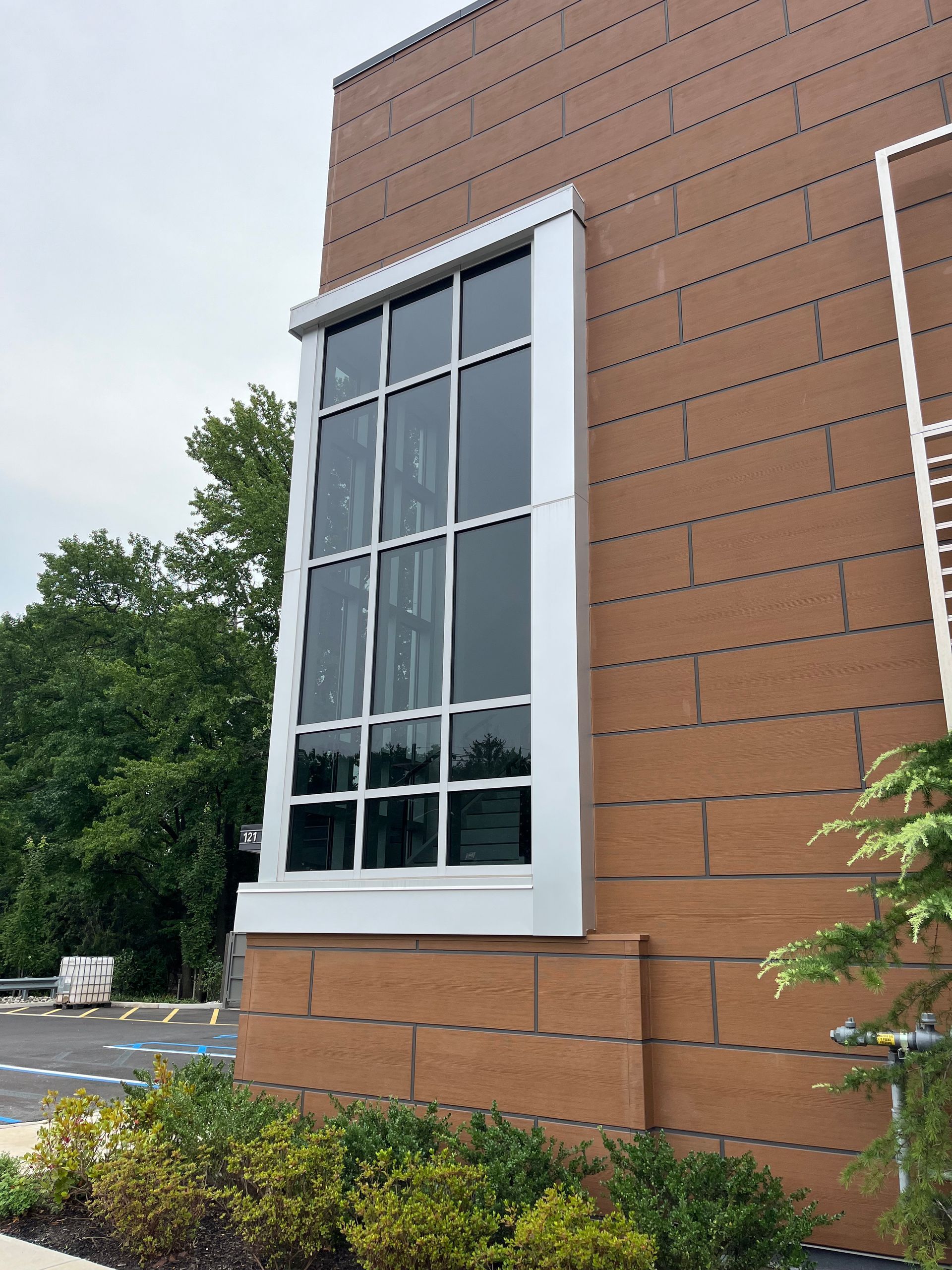 Brick building wall with a tall white-trimmed window beside trees and shrubs