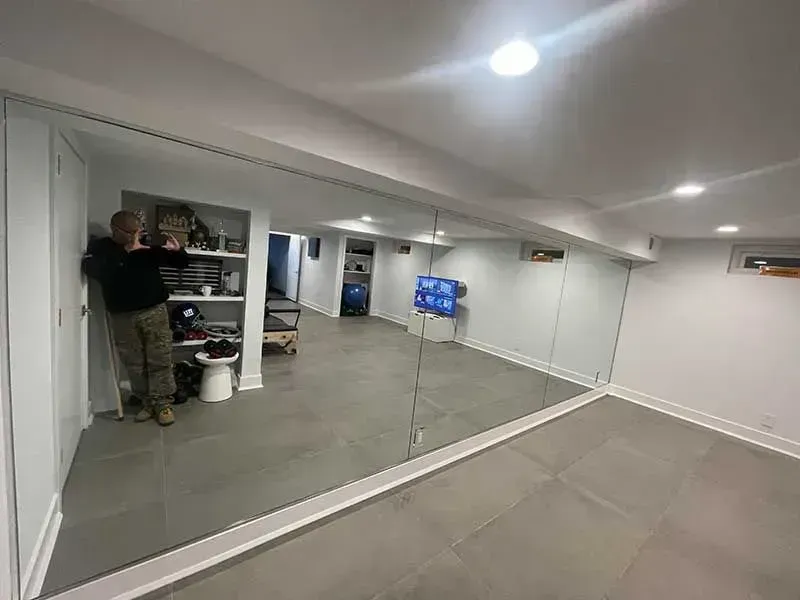 A person stands beside a large wall mirror in a basement room with grey tile floors and recessed lighting.