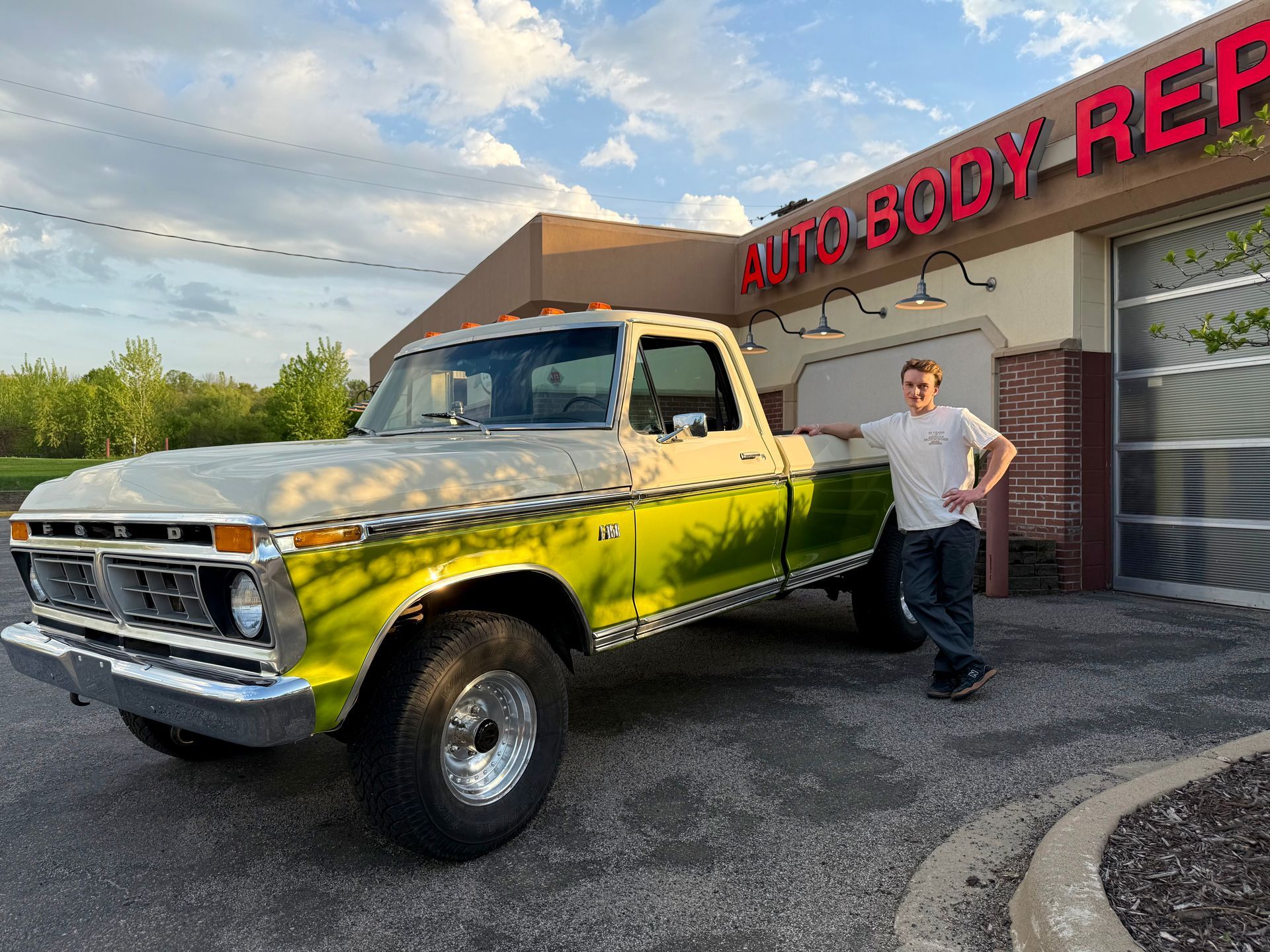 A man is standing next to a green and white truck in front of an auto body shop