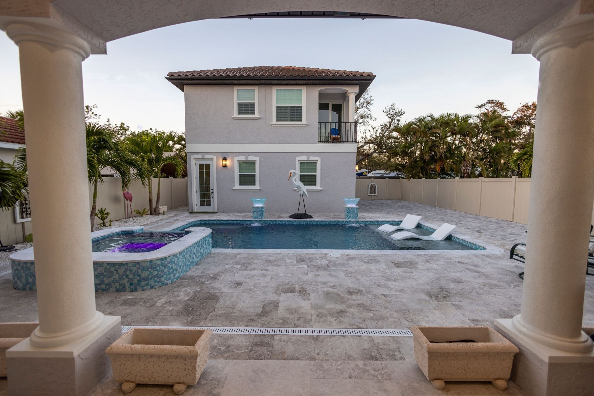 View of a house and pool area with white pillars, blue pool, and lounge chairs.