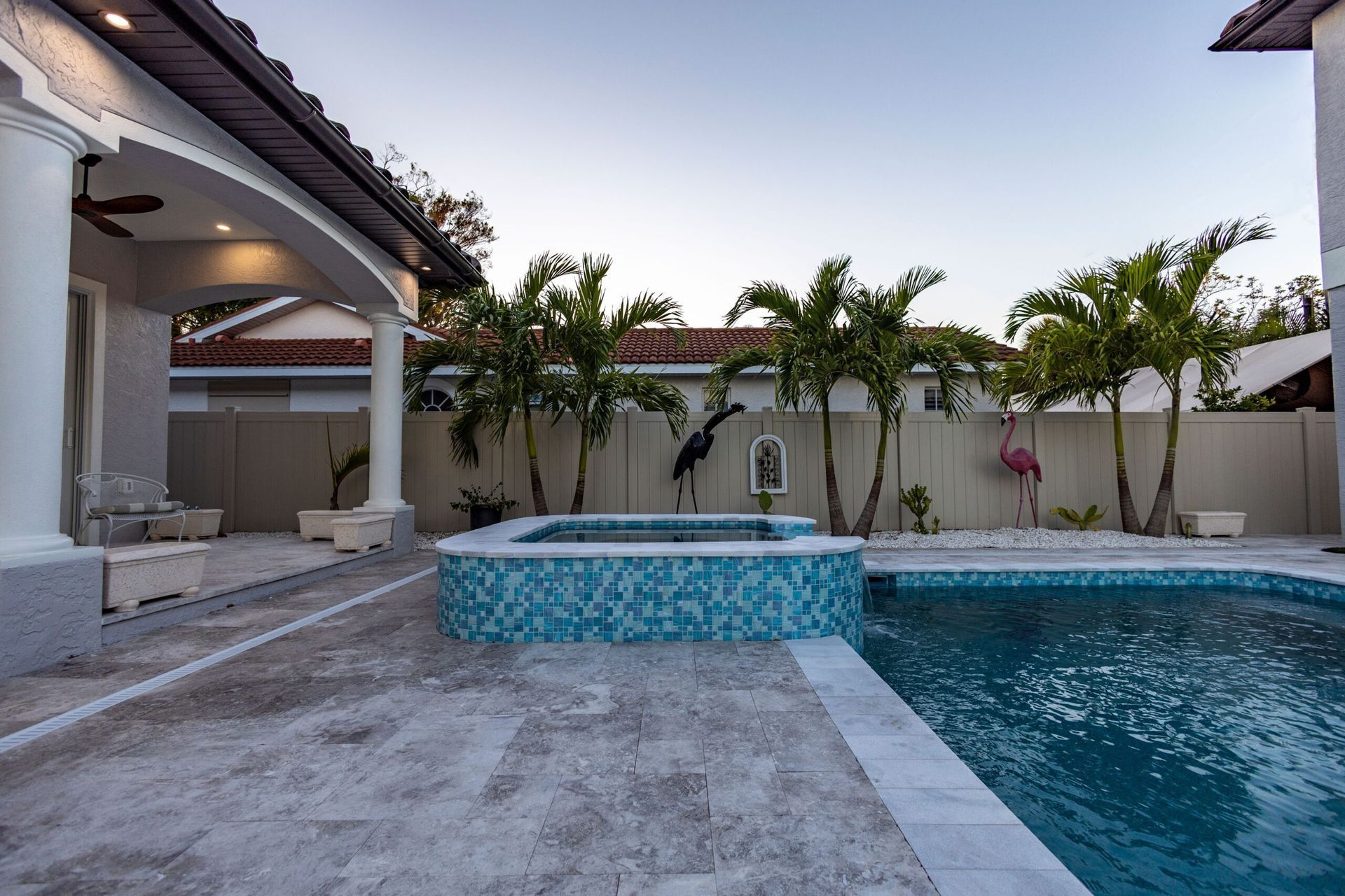 Pool area with turquoise mosaic hot tub, swimming pool, palm trees, and decorative flamingos.