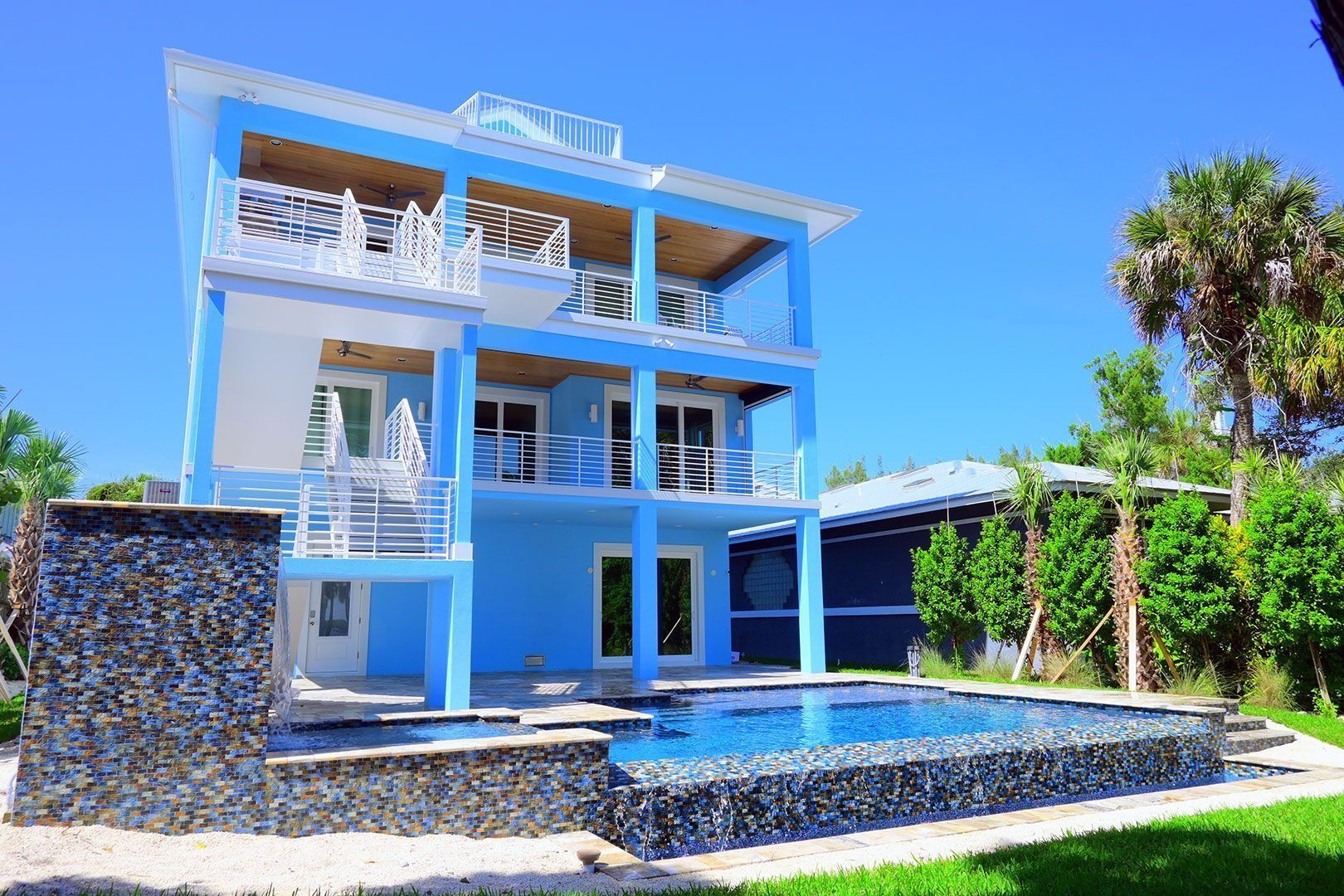 Blue coastal home with balconies, a pool, and palm trees under a sunny sky.