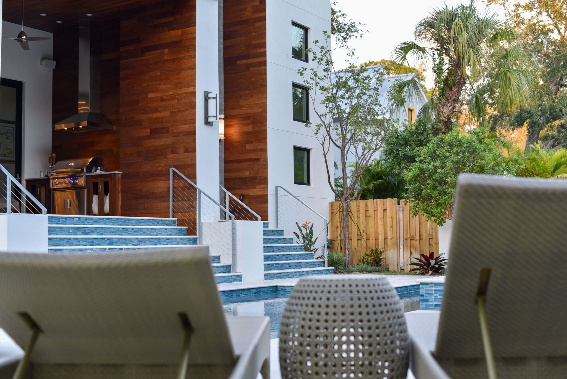 Poolside view of a modern house with a wooden outdoor kitchen and lounge chairs by the water.