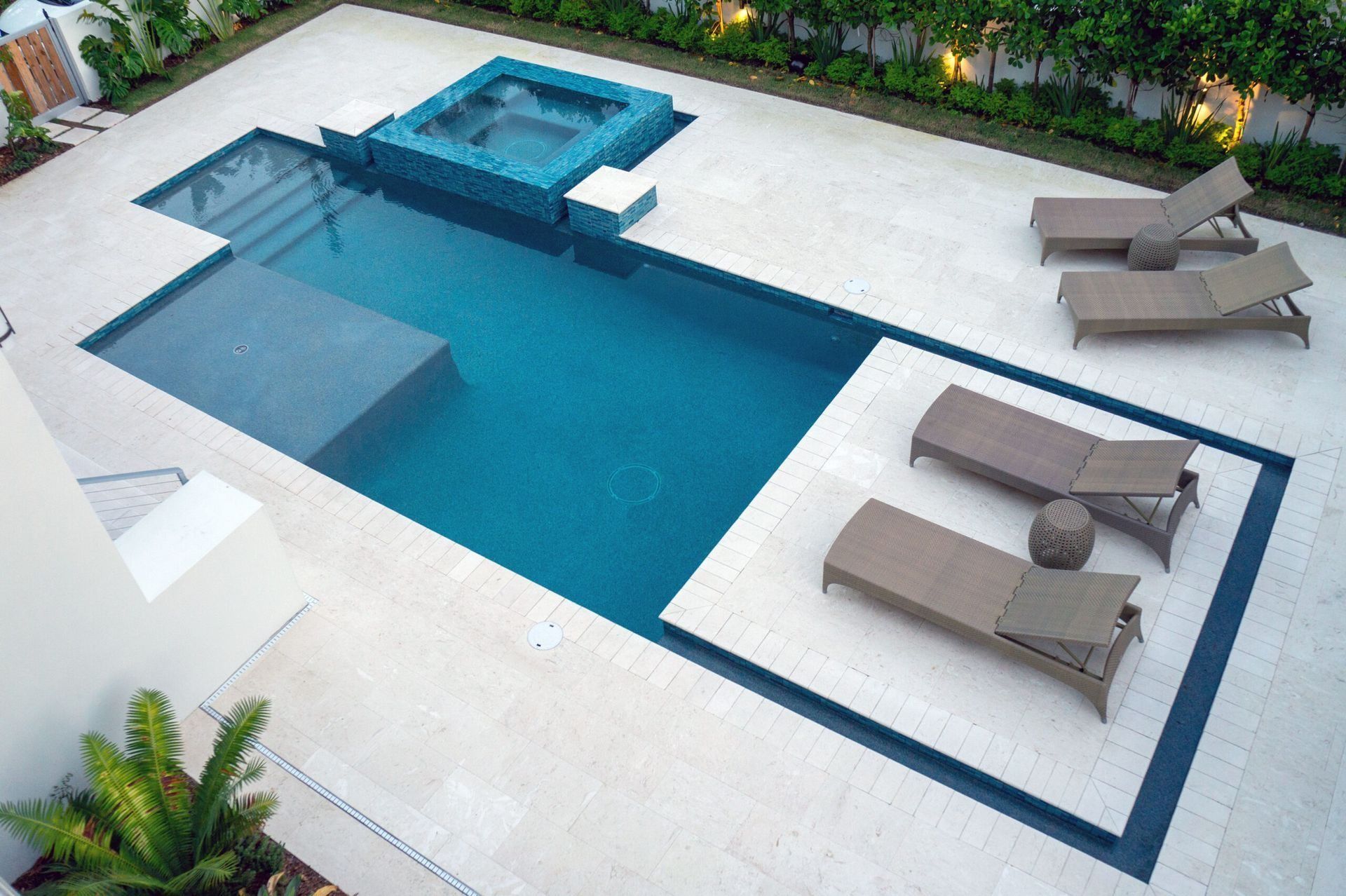 Overhead view of modern turquoise pool with jacuzzi, lounge chairs on light stone patio.