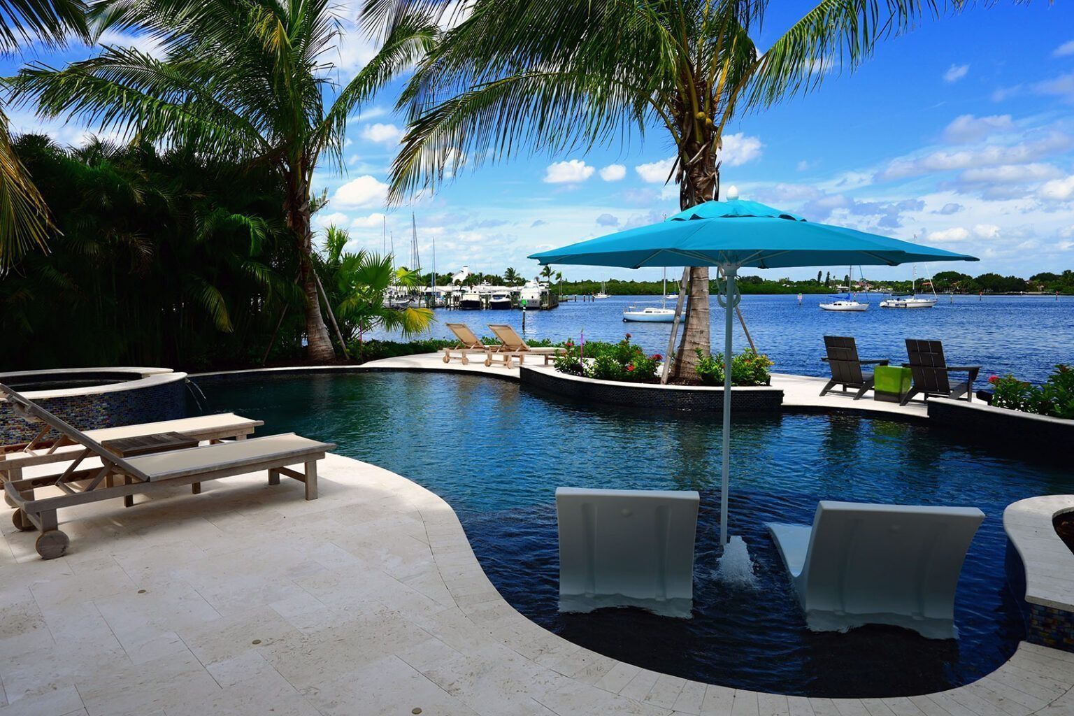 Swimming pool with turquoise umbrella, lounge chairs, palm trees, and water views.