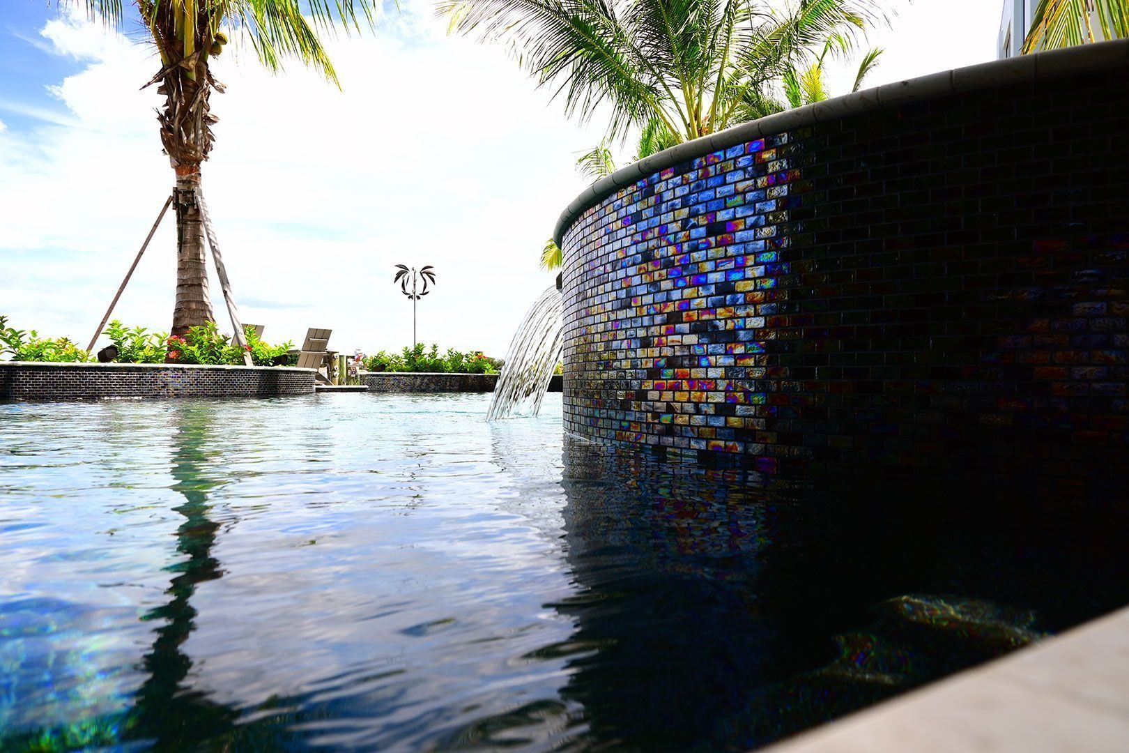 Pool with mosaic tile wall and palm trees under a blue sky. Water gently flows.