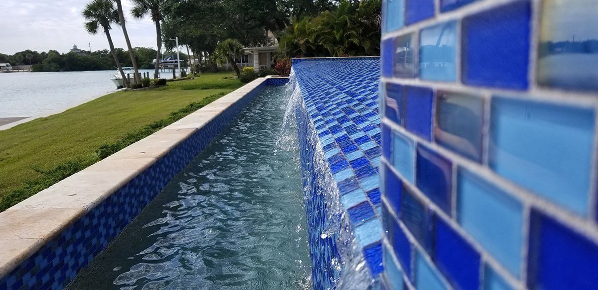 A rectangular swimming pool with blue tiles and cascading water; background includes a waterfront and green grass.