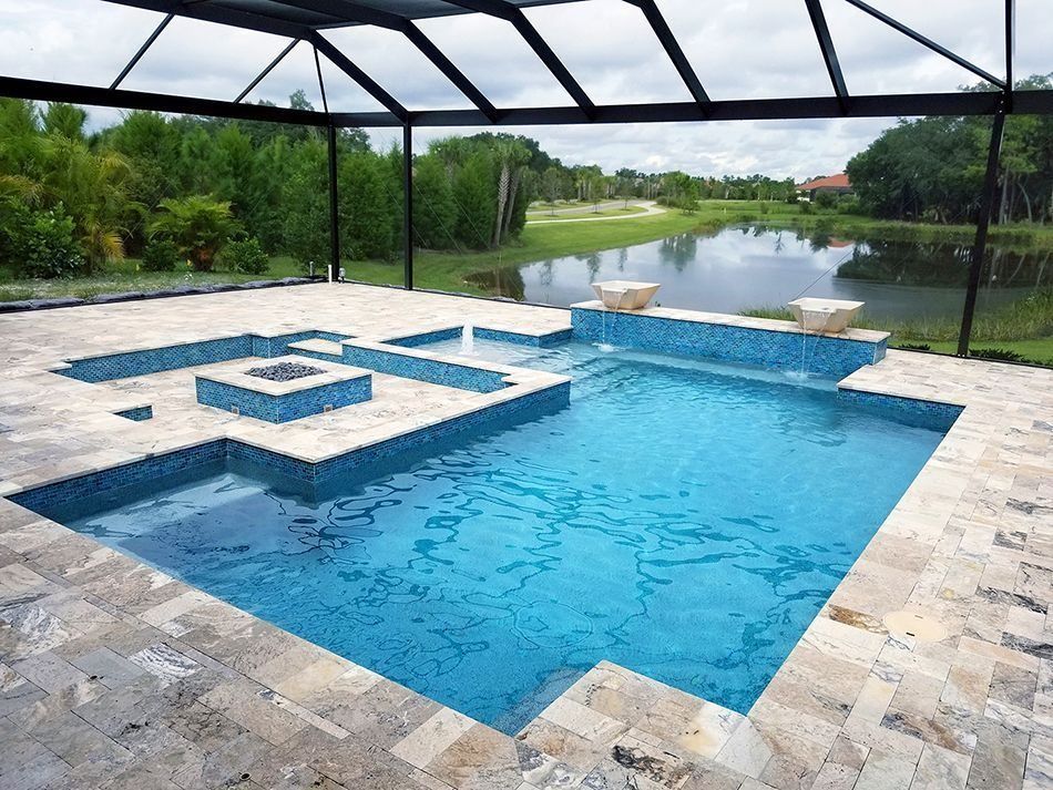 A blue tiled swimming pool with a unique geometric design, surrounded by stone paving and a screened enclosure, overlooking a lake.
