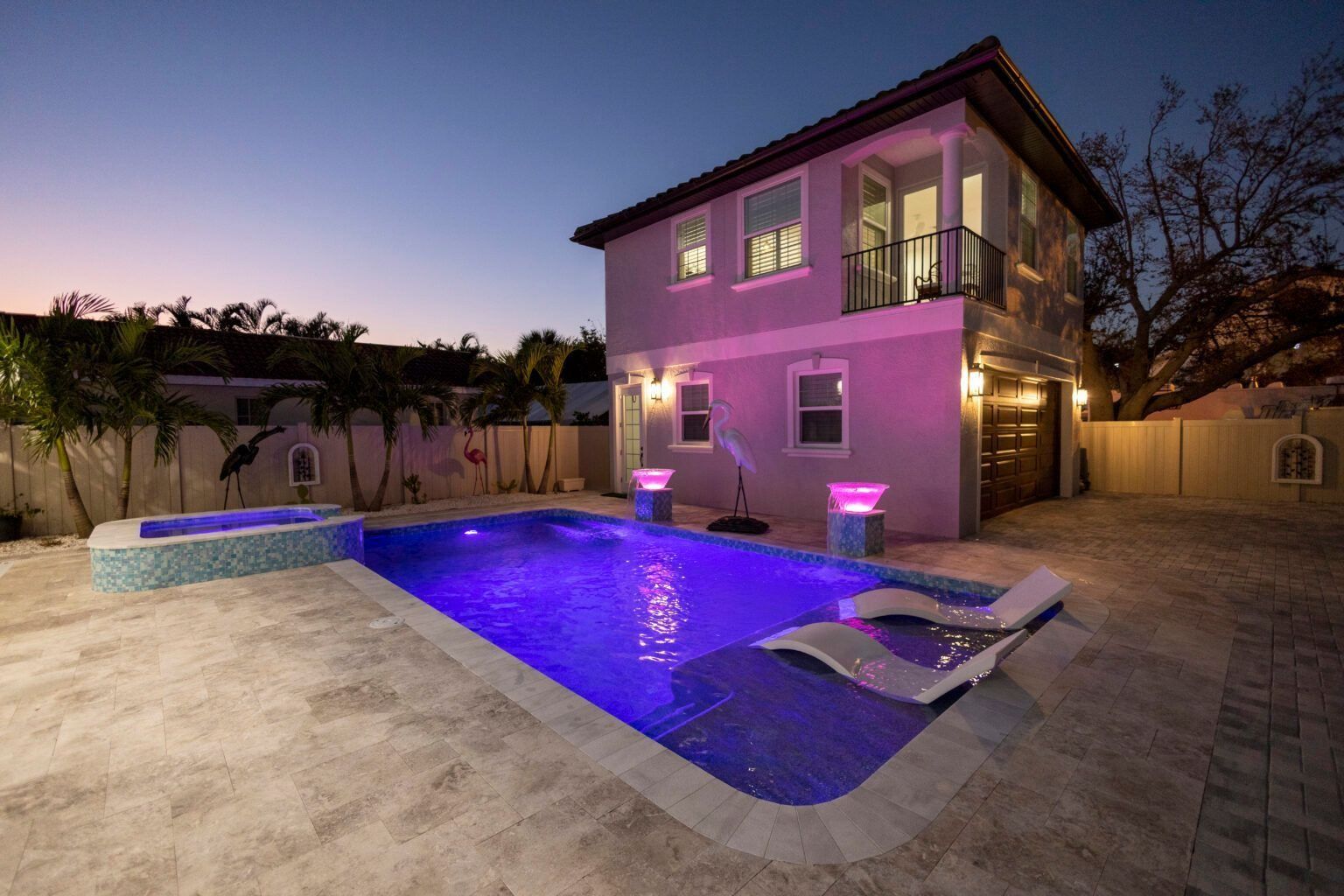 Night view of a house with a lit pool and jacuzzi. Purple lights illuminate the pool and building. Lounge chairs on the patio.