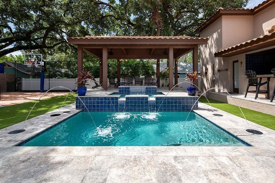 Swimming pool with water fountains, a shaded patio, and a basketball hoop in a backyard.