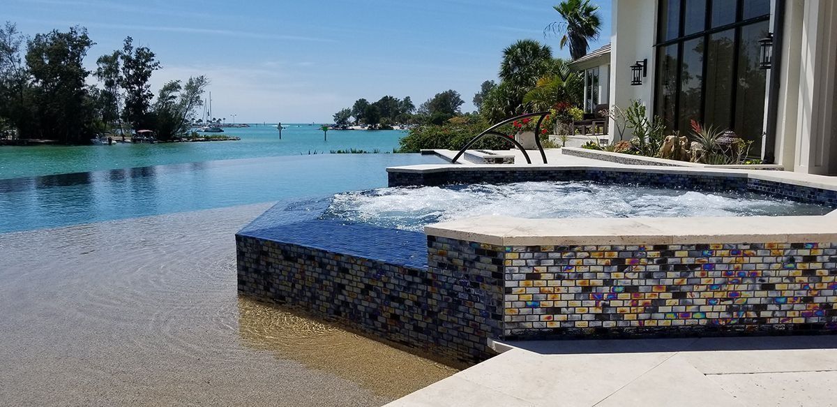 A pool with an infinity edge, overlooking a turquoise body of water. A jacuzzi with bubbling water is in the foreground.