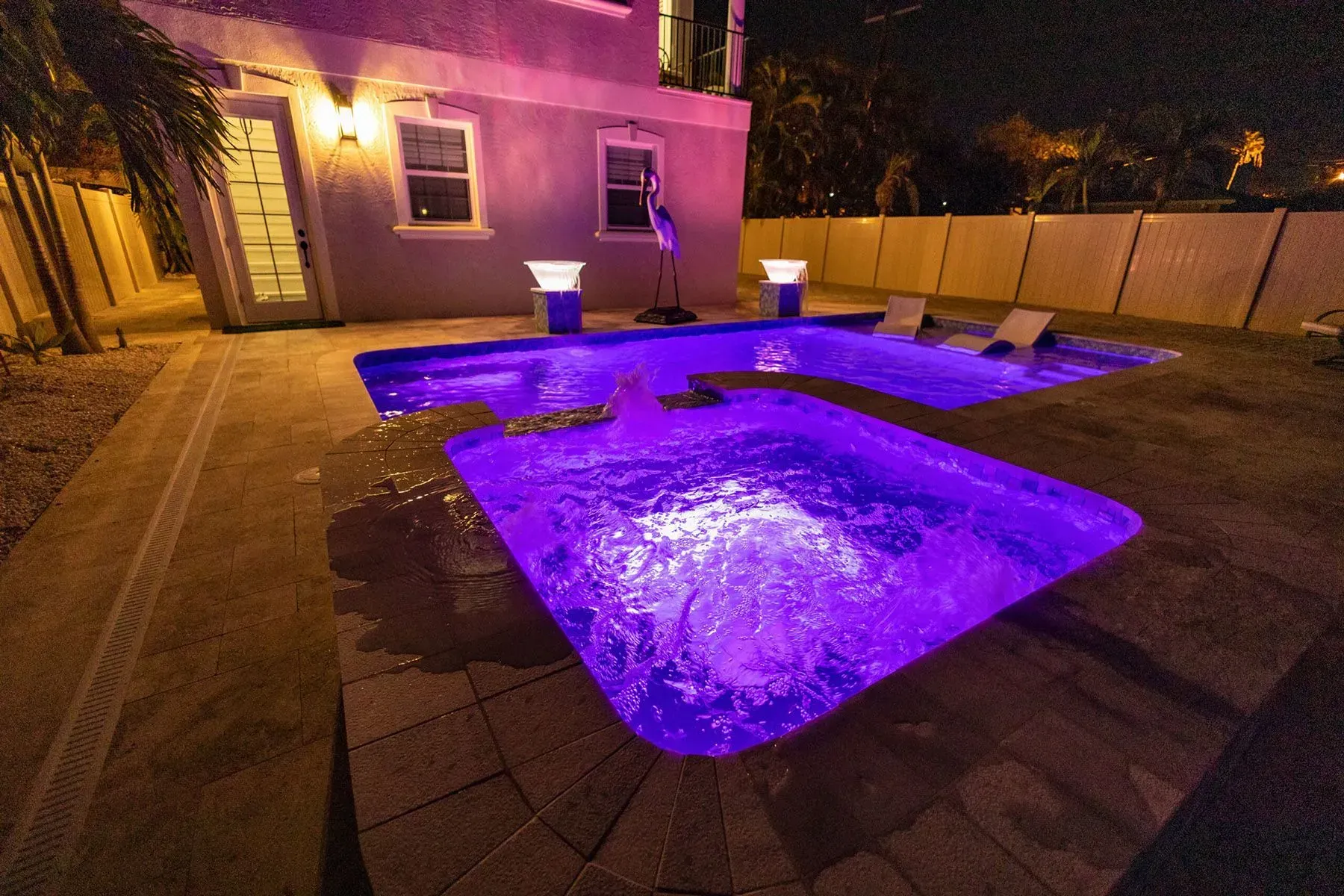 Nighttime backyard with purple-lit pool, hot tub, lounge chairs, and house.