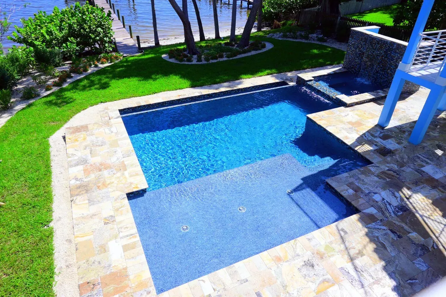 Blue-tiled swimming pool with steps, surrounded by stone patio and grass, with a view of the water.