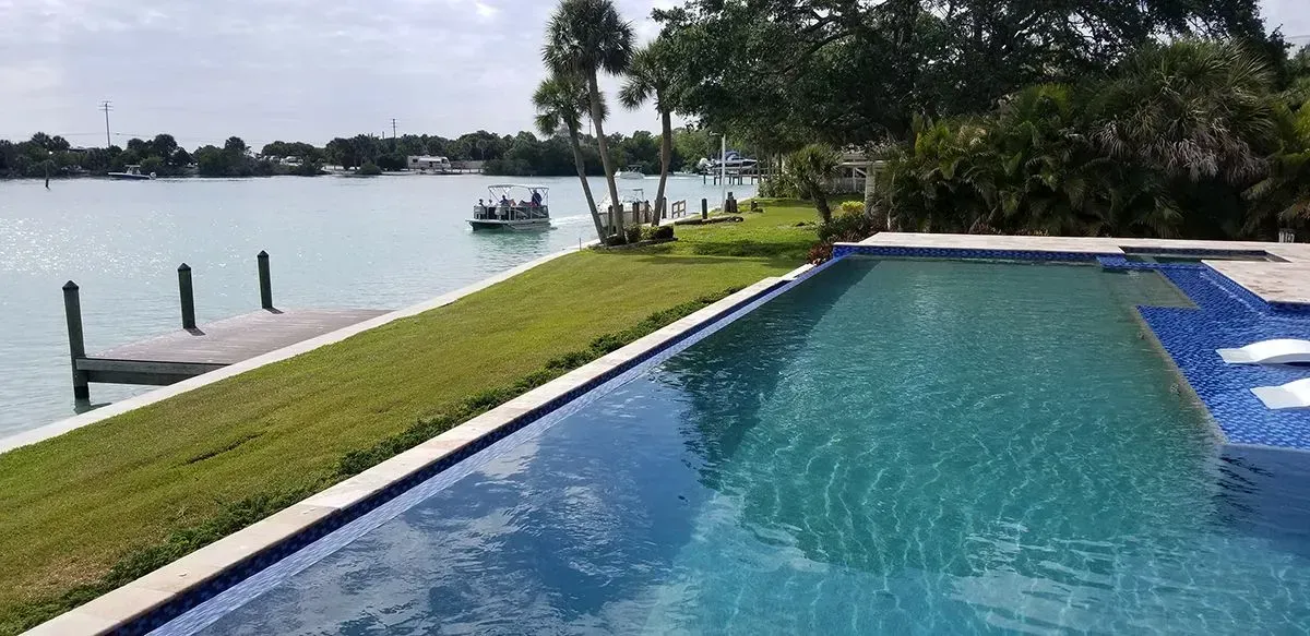 A waterfront view featuring a pool, a boat on the water, and a dock. Green grass lines the edge.