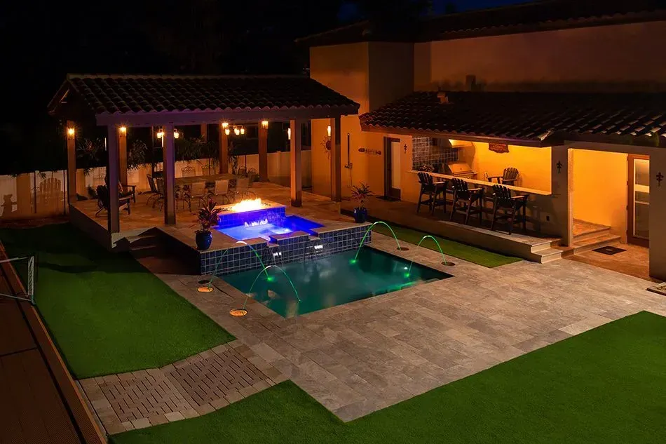 Night view of a luxurious backyard with a pool, fire pit, and outdoor kitchen illuminated by warm lighting.