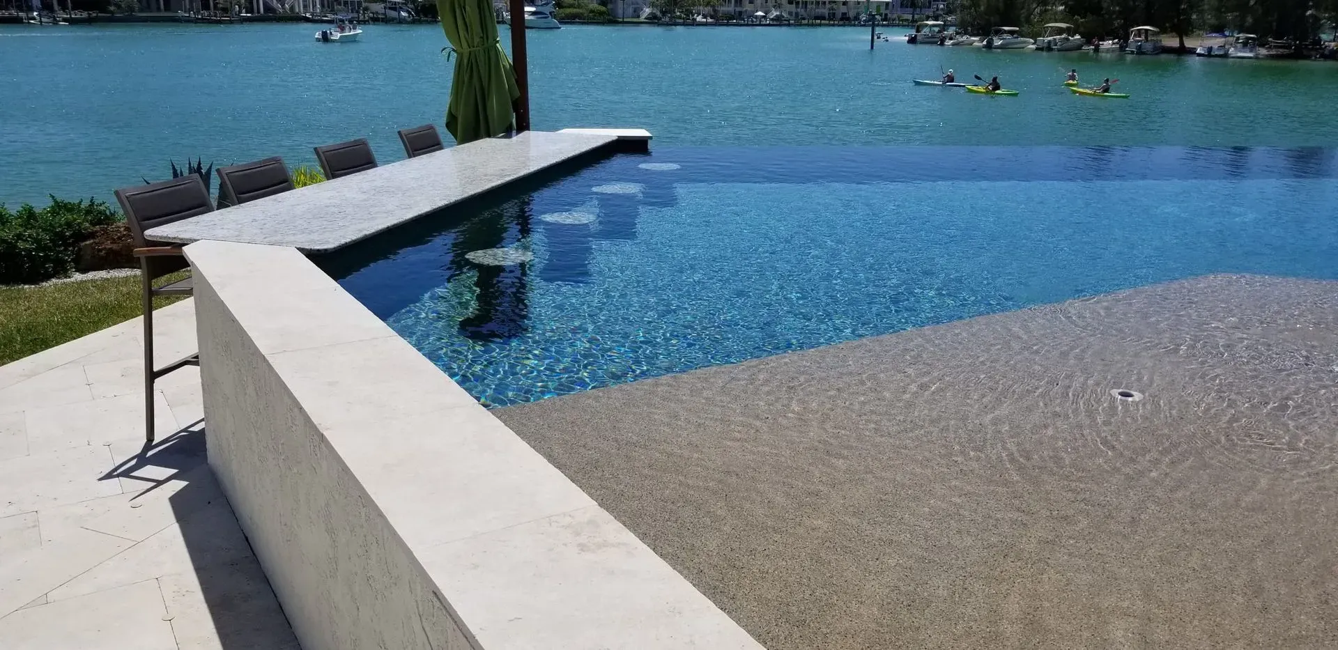 An infinity pool with blue water overlooking a lake. Light colored deck and wall with seating.