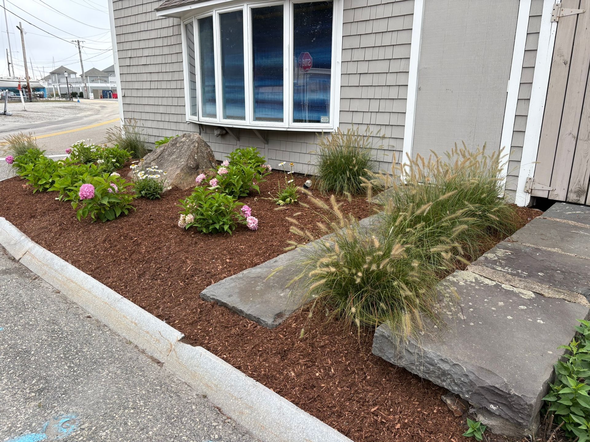 Flower bed with pink blooms, a large rock, and ornamental grass near a building.