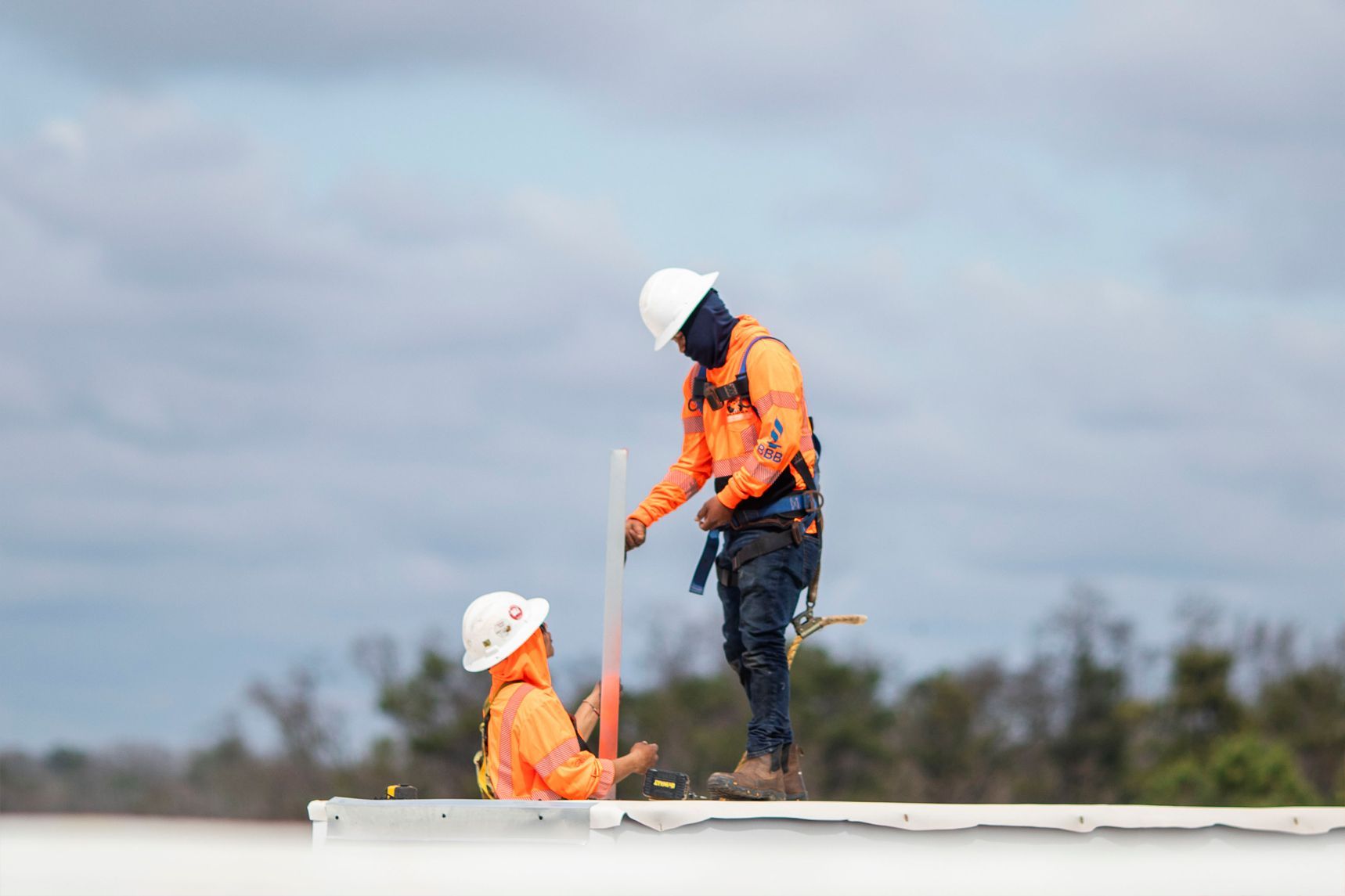 Two workers in orange safety gear and white hard hats on a rooftop under a cloudy sky