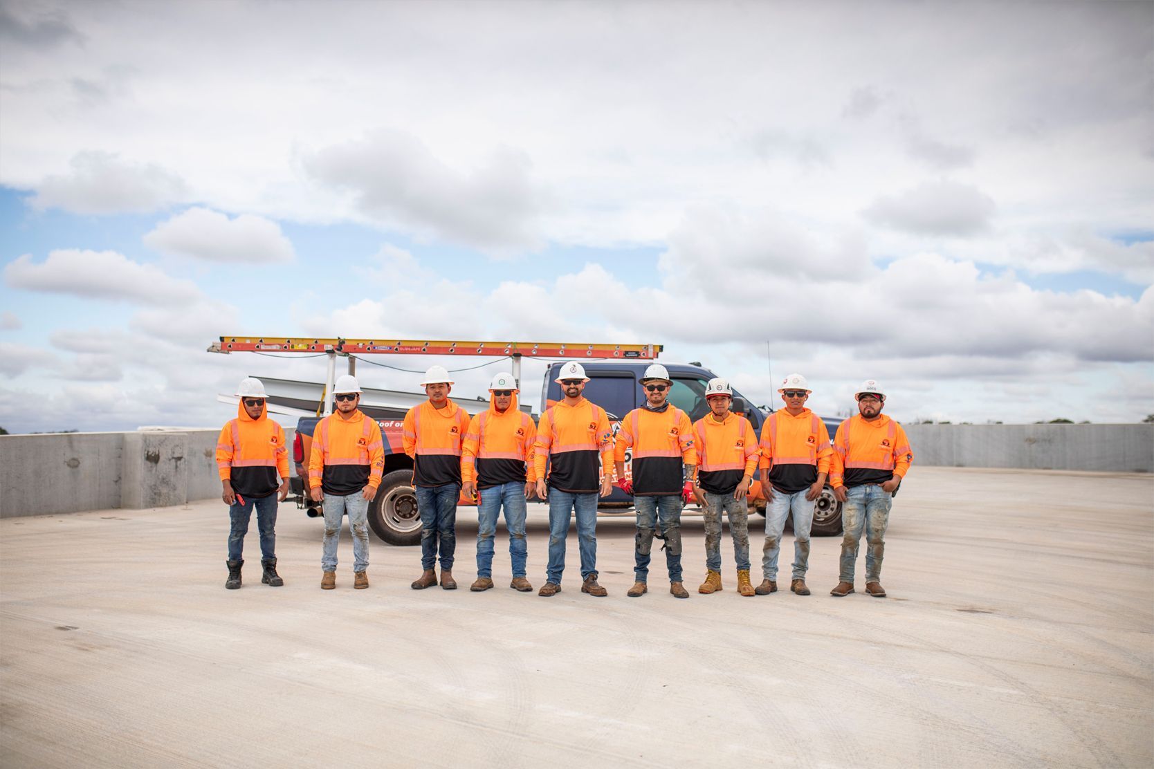 Group of workers in orange safety shirts and hard hats standing on a construction site under a cloudy sky